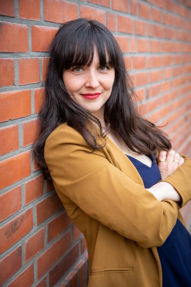 Frau mit langen, dunklen Haaren in blauer Bluse und braunem Blazer, lächelt vor Ziegelwand.