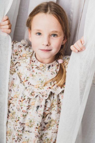 Mädchen mit Zopf in einem blumigen Kleid, schaut durch einen Vorhang.