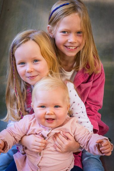 Drei Mädchen posieren fröhlich, die älteren lachend hinter dem jüngeren Baby.