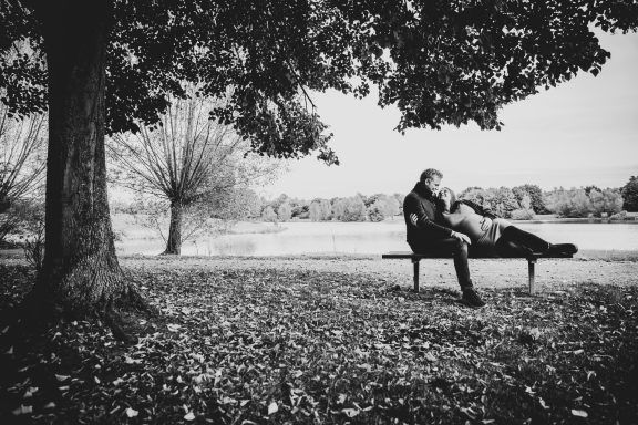 Zwei Personen sitzen auf einer Bank unter einem Baum in einer herbstlichen Landschaft.