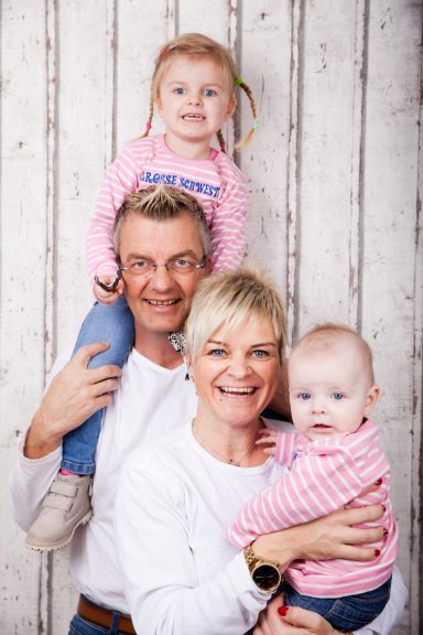 Familienporträt: Ein Paar mit zwei Kleinkindern, alle lächeln vor einem hellen Hintergrund.