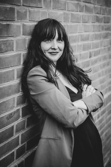 Frau mit langen Haaren und Blazer lächelt vor einer Backsteinmauer.