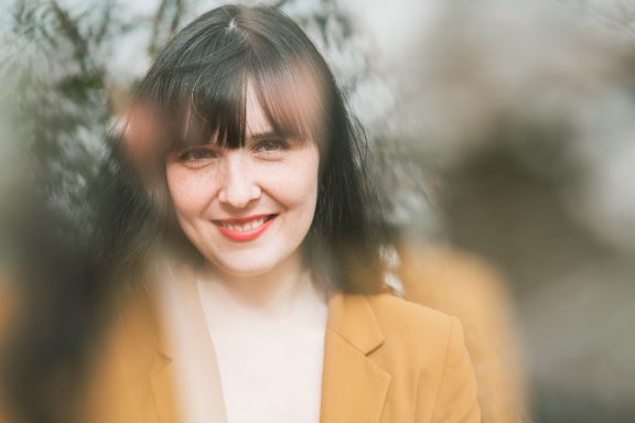 Eine lächelnde Frau mit langen, dunklen Haaren, trägt einen hellbraunen Blazer.