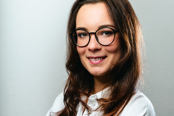 Frau mit Brille und langen, dunklen Haaren, lächelt freundlich vor einem grauen Hintergrund.