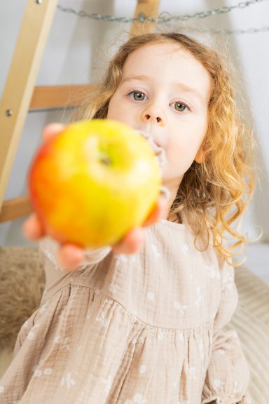 Kleines Mädchen mit lockigem Haar hält einen roten Apfel in die Kamera.
