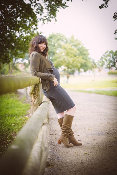 Schwangere Frau in Stiefeln lehnt an einem Holzgeländer in einer grünen Landschaft.