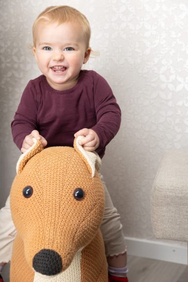 Ein lächelndes Kleinkind sitzt auf einem Stofftier-Fuchs und trägt einen burgunderfarbenen Pullover.