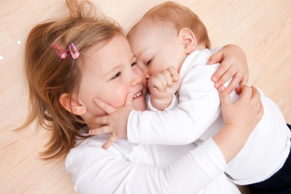 Ein kleines Mädchen umarmt ein Baby, beide lächeln fröhlich auf dem Boden.