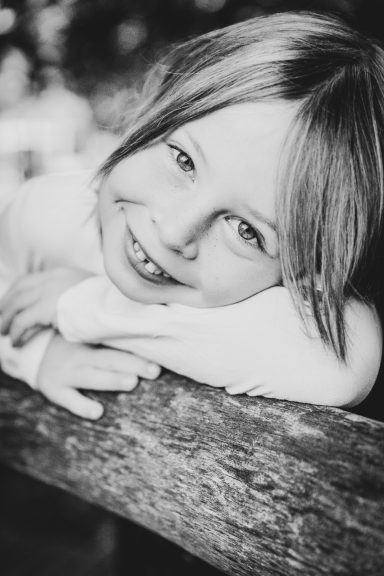 Lächelndes Kind mit blonden Haaren, die auf einem Holzbalken lehnt.