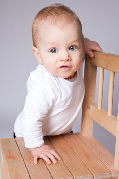 Ein Baby mit blauen Augen lehnt sich an einen Holzstuhl und schaut neugierig.