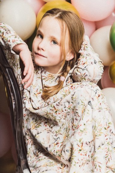 Ein Mädchen mit Zopf sitzt in einem gemusterten Kleid vor bunten Luftballons.