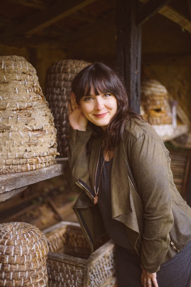 Frau mit dunklen Haaren lehnt an einer Wand aus Weidenkörben und lächelt.