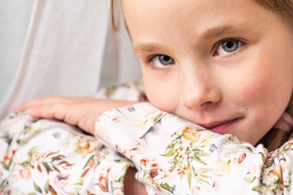 Ein Mädchen mit blonden Haaren, lächelnd und in einem blumigen Hemd.