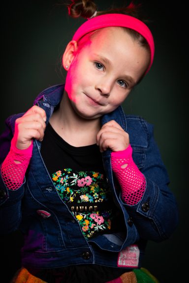 Ein Mädchen mit einem blauen Jeansjackett und pinkem Haarband, lächelt fröhlich.