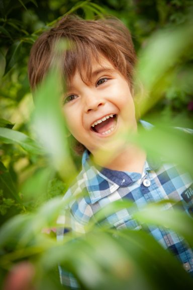 Ein lächelnder Junge mit braunen Haaren, der durch Pflanzen blickt.