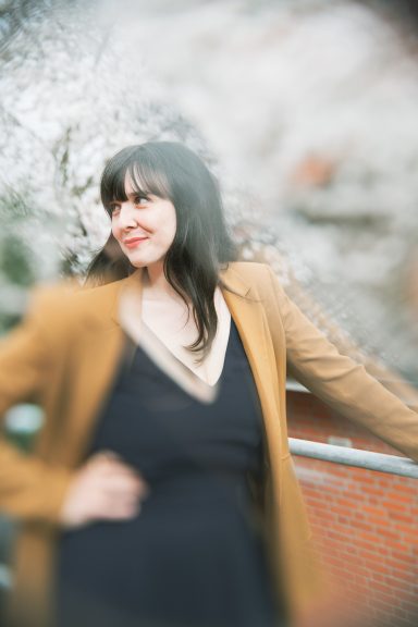 Frau mit dunklen Haaren in einem braunen Mantel vor blühenden Bäumen.