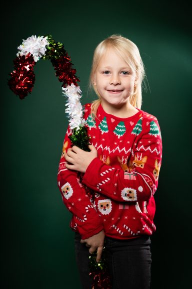 Mädchen mit einem Weihnachtsbaum-Sweater hält einen mit Glitzer verzierten Zuckerstangen-Stab.