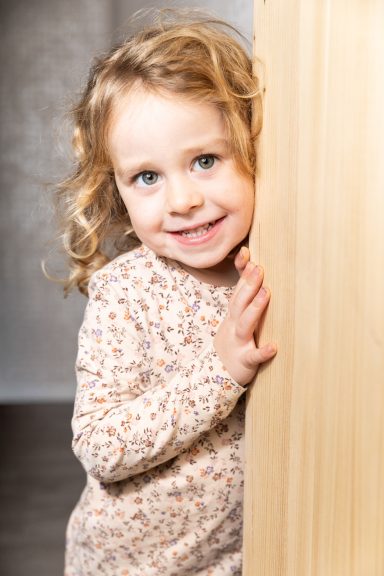 Lächelndes Mädchen mit lockigem Haar, das an einem Holzmöbel steht.