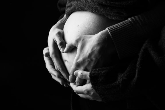 Hände um einen schwangeren Bauch, in Schwarz-Weiß gehalten.