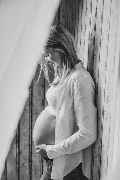 Schwangere Frau mit erhobenem Bauch, lehnt an einer Holzwand und lächelt.