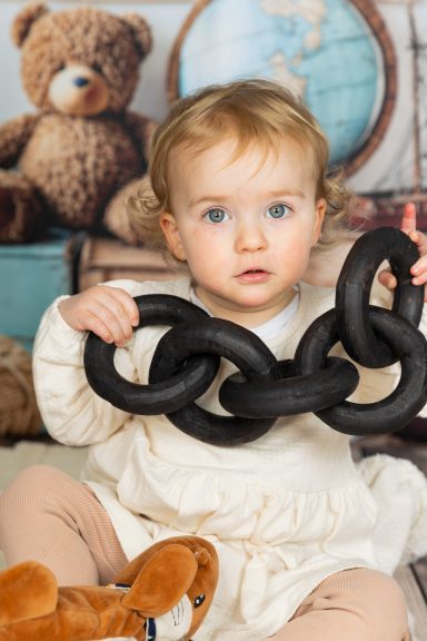 Ein Kleinkind sitzt und hält eine große schwarze Kette aus Spielzeug. Im Hintergrund sind Spielzeuge.