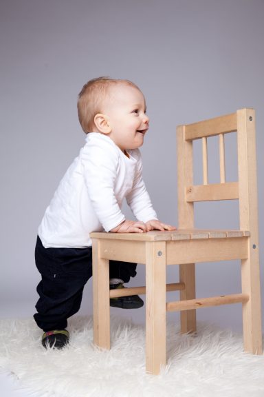 Ein lächelndes Baby steht neben einem kleinen Holzstuhl.