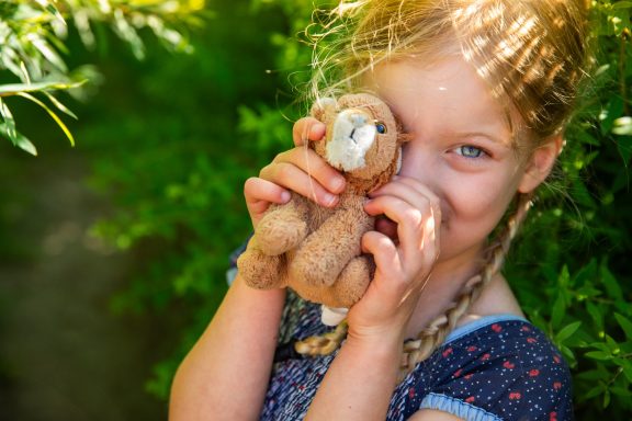Mädchen mit Zöpfen hält ein Plüschbärchen und lächelt schelmisch.