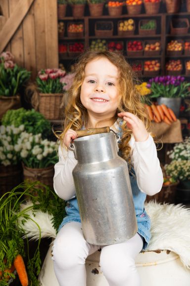 Kleines Mädchen mit lockigem Haar hält einen Milchkann neben einem Gemüse- und Blumenstand.