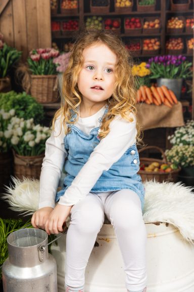 Ein kleines Mädchen mit lockigem Haar sitzt auf einem Hocker in einem Marktsetting.