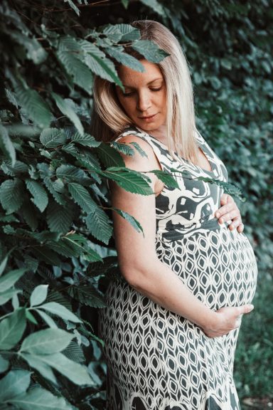 Schwangere Frau in einem gemusterten Kleid, umgeben von grünen Blättern.