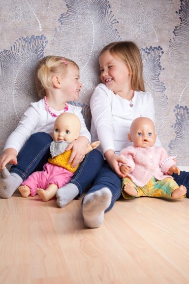 Zwei Mädchen sitzen mit Puppen auf dem Boden und schauen sich lächelnd an.