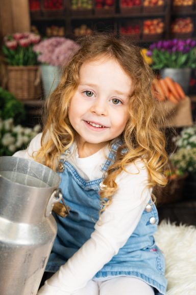 Lächelndes Kind mit lockigem Haar, das einen Milchkübel hält, umgeben von Blumen und Gemüse.