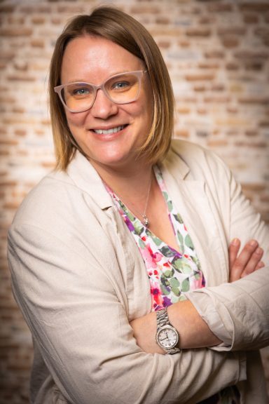 Lächelnde Frau mit Brille, die die Arme verschränkt, vor einer Backsteinwand.