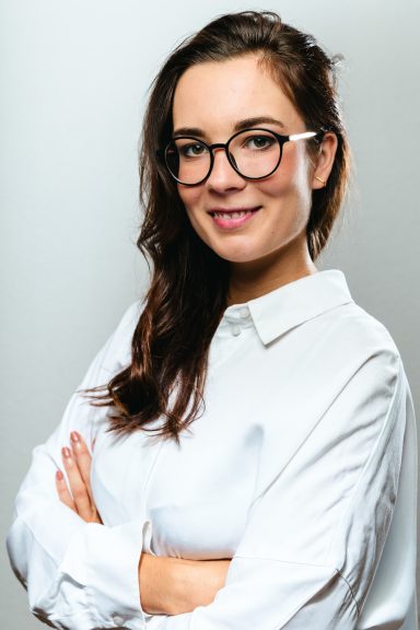 Junge Frau mit Brille und weißem Hemd, lächelt selbstbewusst.