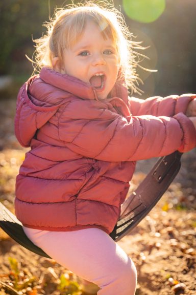 Kleines Mädchen in einer Sonnenstrahlen mit einer pinken Jacke, das auf einer Schaukel sitzt.