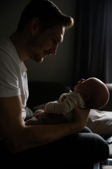 Vater hält sein Neugeborenes, beide schauen sich liebevoll an. Dämmerlicht im Hintergrund.