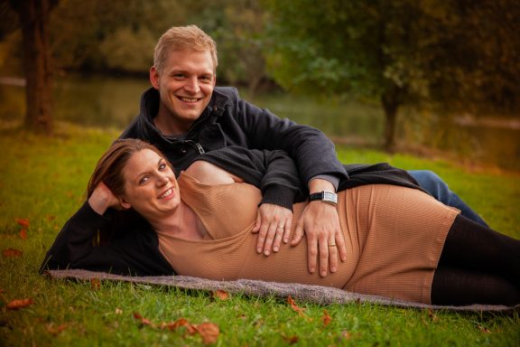 Paar liegt auf einer Decke im Freien, umgeben von Natur und Wasser, lächelnd.
