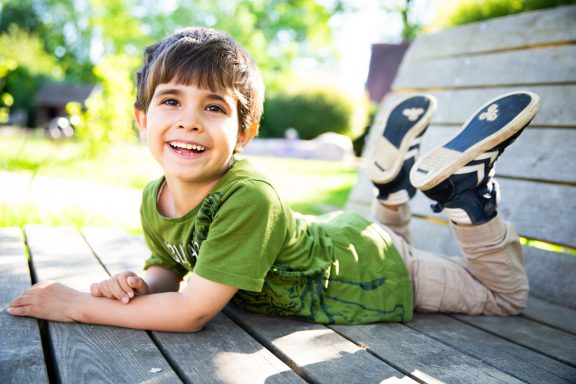 Ein Junge lächelt entspannt auf einer Holzbank, barfuß mit einem grünen T-Shirt.