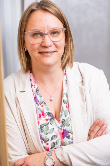 Frau mit Brille, lächelnd, in leichter Kleidung mit floralem Muster, Arme verschränkt.