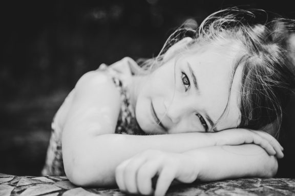 Ein Mädchen mit langen, hellen Haaren lehnt sich nachdenklich auf einen Tisch.