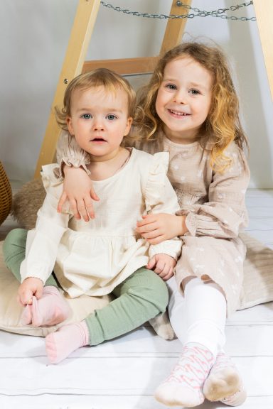 Zwei kleine Mädchen sitzen zusammen, eines trägt einen hellen Pullover, das andere ein Kleid.
