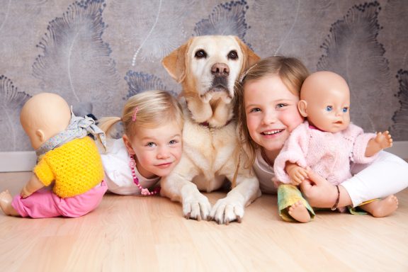 Zwei Kinder mit Puppen und einem Hund auf einem Holzfußboden.