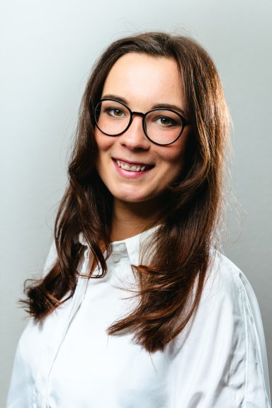 Junge Frau mit langen, braunen Haaren und Brille, lächelt in einem hellen Oberteil.