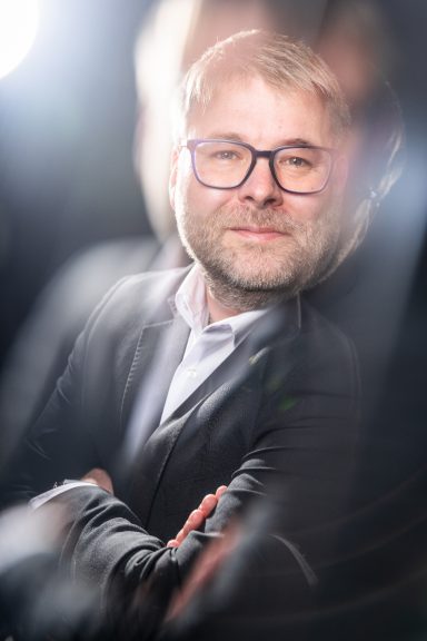 Mann mit Brille und kurzem Haar, lächelnd, in formeller Bekleidung.