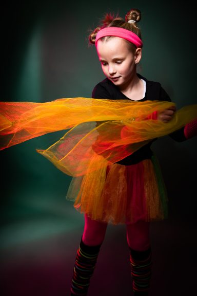 Ein kleines Mädchen in einem bunten Tutu hält einen farbigen Schal und lächelt.