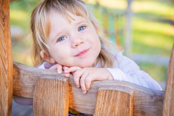 Ein kleines Mädchen mit blonden Haaren lächelt und stützt den Kopf auf eine Holzbank.