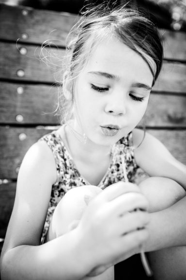 Mädchen sitzt auf einer Bank und pustet einen Wunsch in die Luft.