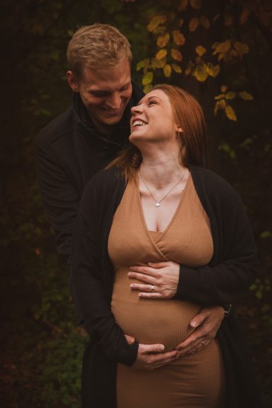 Schwangere Frau in Umarmung mit Partner, beide lächeln glücklich. herbstliche Atmosphäre.