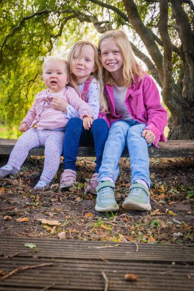 Drei Kinder sitzen lächelnd im Freien auf einer Holzbank unter Bäumen.