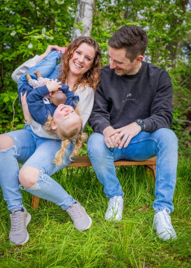 Glückliche Familie im Freien: Mutter hält lachendes Kind, Vater sitzt daneben.